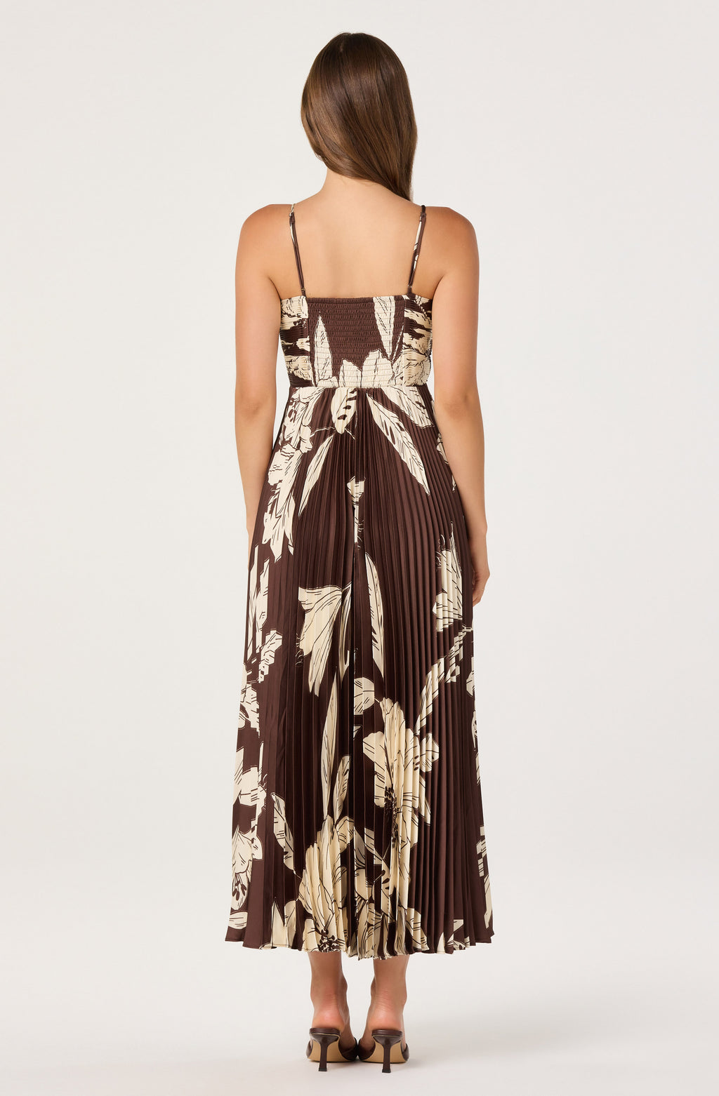 A woman viewed from the back wearing a sleeveless pleated maxi dress with thin straps and a brown and beige floral print, paired with brown heels, against a light gray studio background.