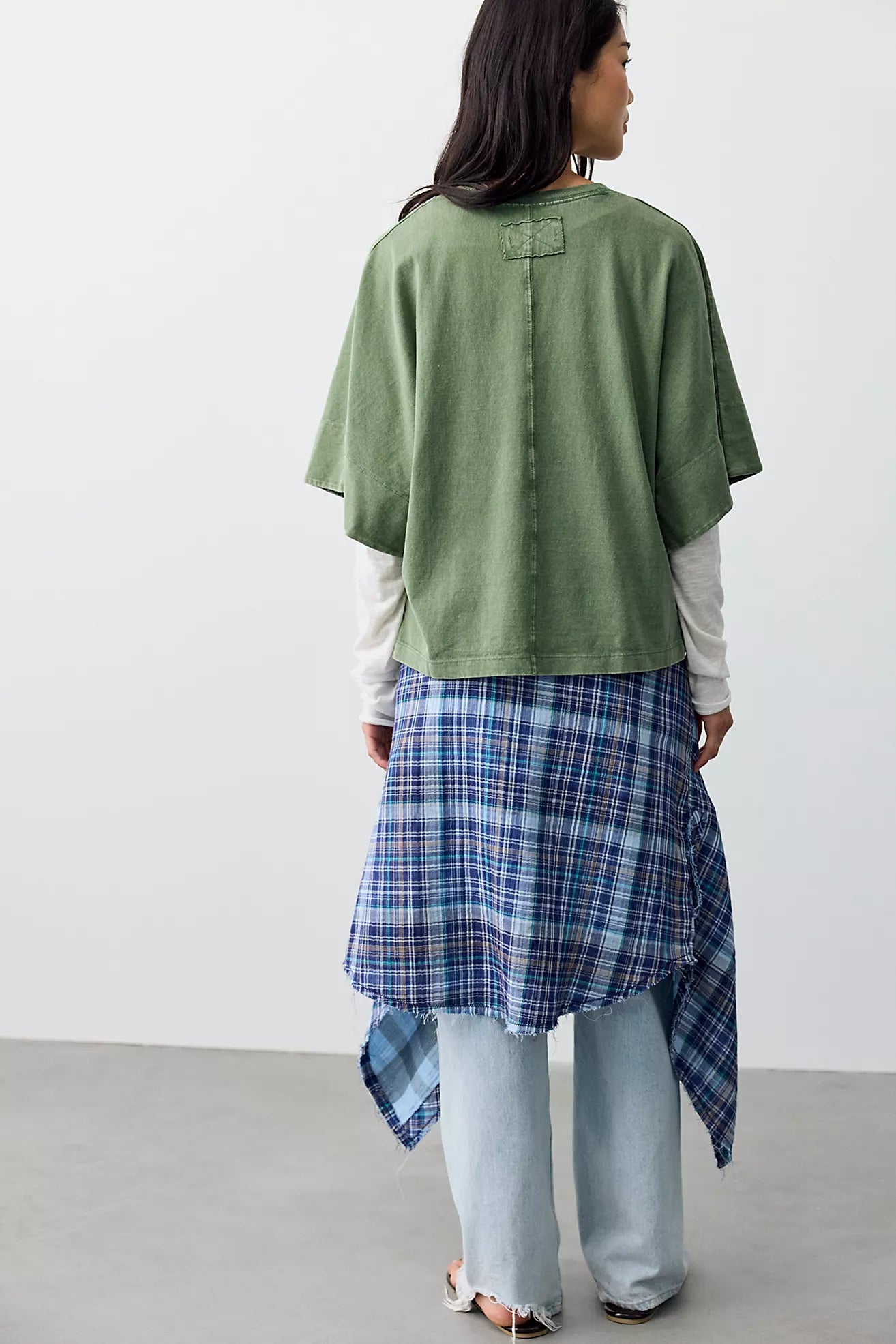 A woman shown from the back wearing a sage green short-sleeve top over a white long-sleeve shirt, paired with a blue and white plaid asymmetrical skirt and light-wash jeans.