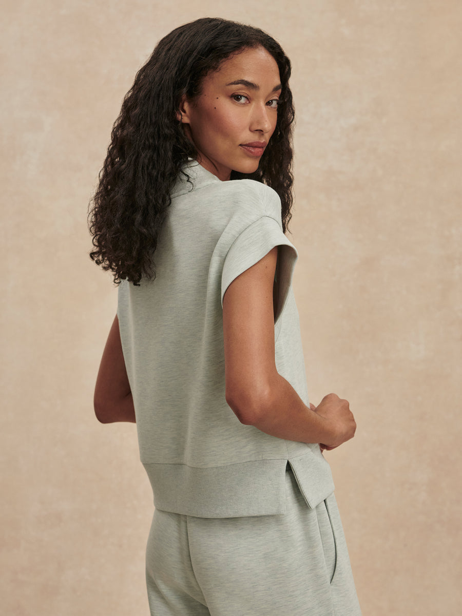 A woman with long, curly dark hair is shown from the back wearing a light sage green short-sleeve knit top and matching sweatpants against a textured beige studio backdrop.