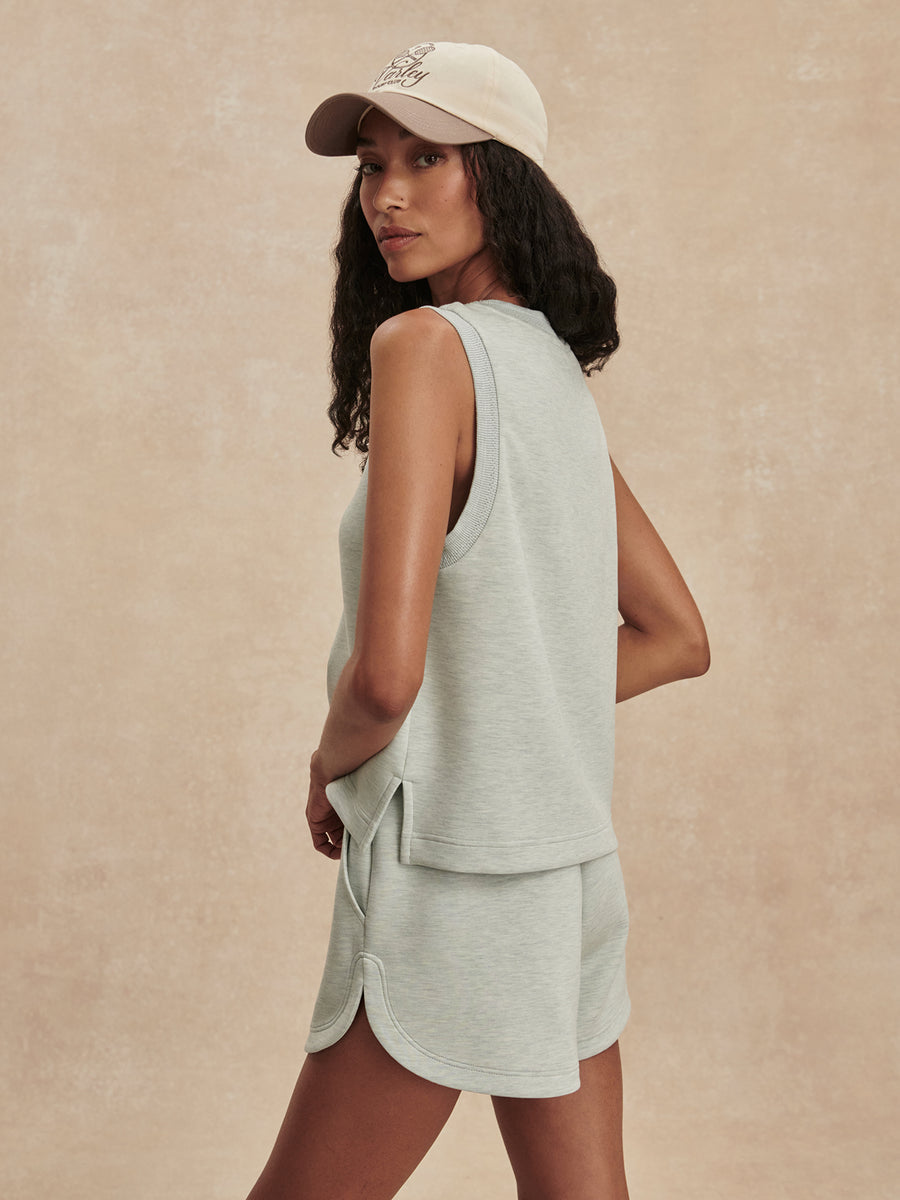 A woman wearing a light sage green sleeveless top and matching shorts, paired with a beige baseball cap, photographed against a textured beige studio backdrop.