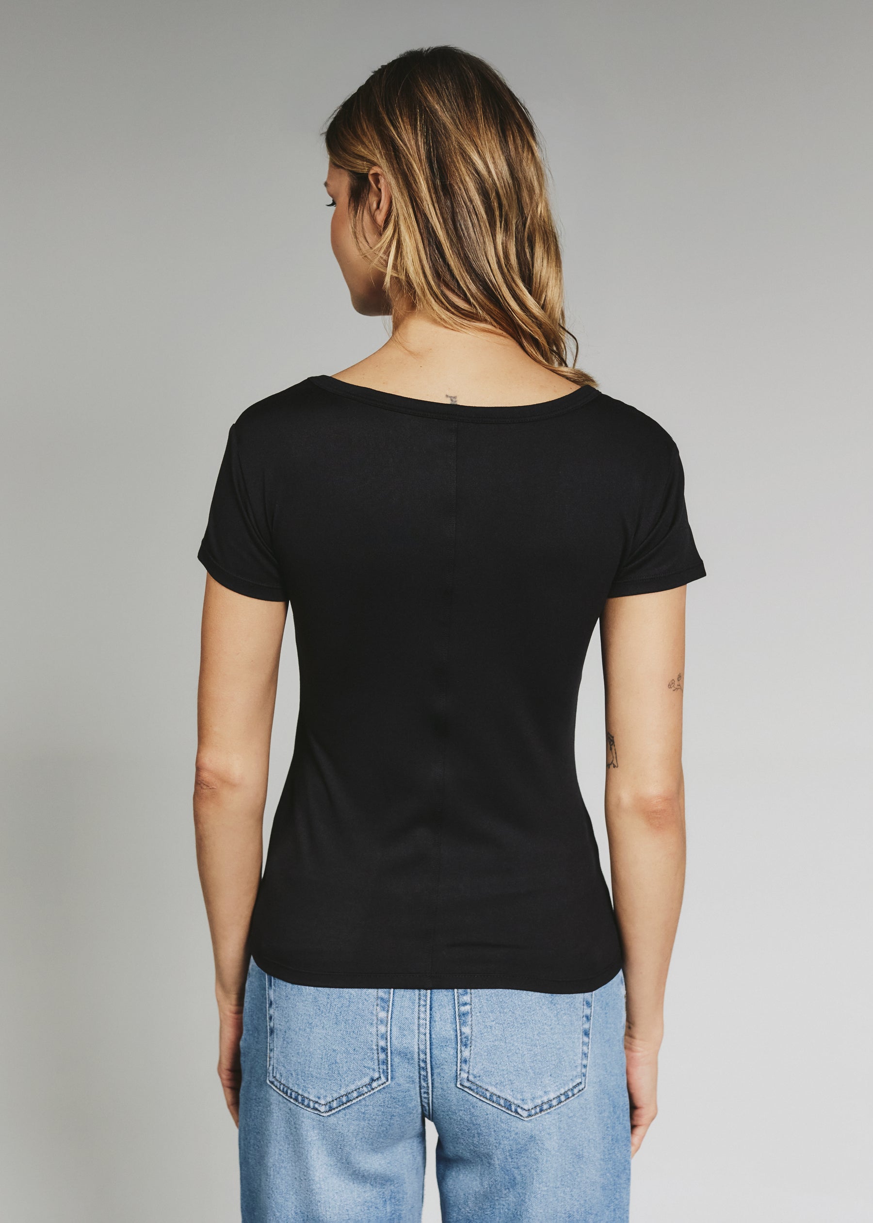 A woman viewed from the back wearing a black short-sleeve t-shirt and light blue jeans, standing against a neutral gray studio background.