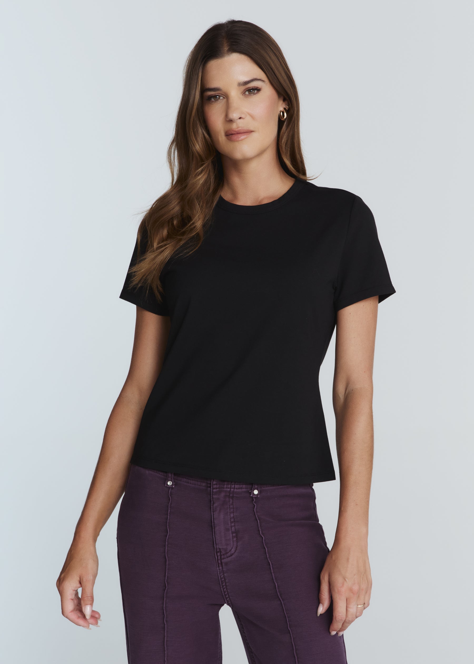 A woman modeling a black short-sleeve crewneck t-shirt paired with dark purple pants, set against a solid light gray studio background.