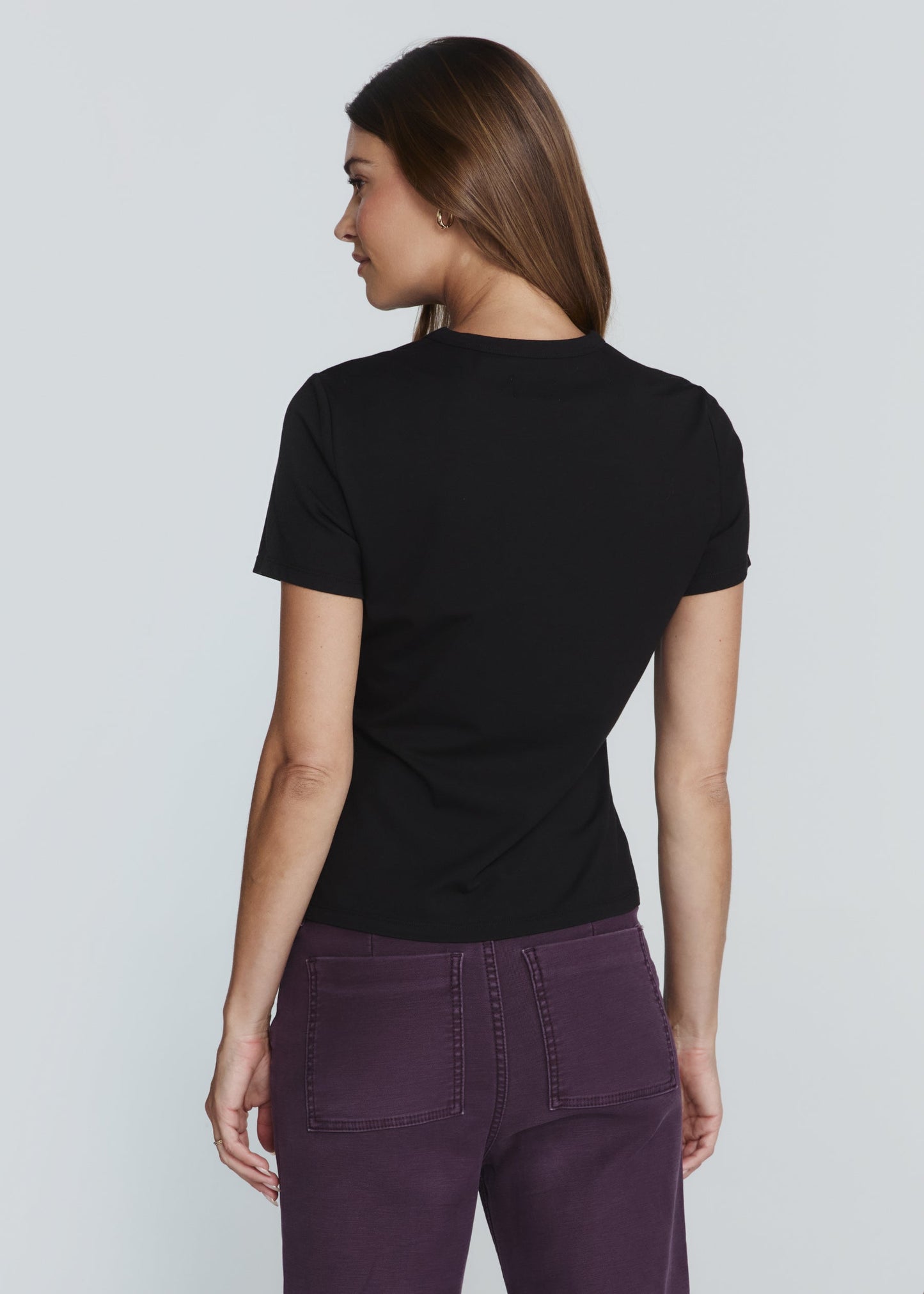 A woman shown from the back wearing a black short-sleeve crewneck t-shirt and purple pants, photographed against a solid light gray studio background.