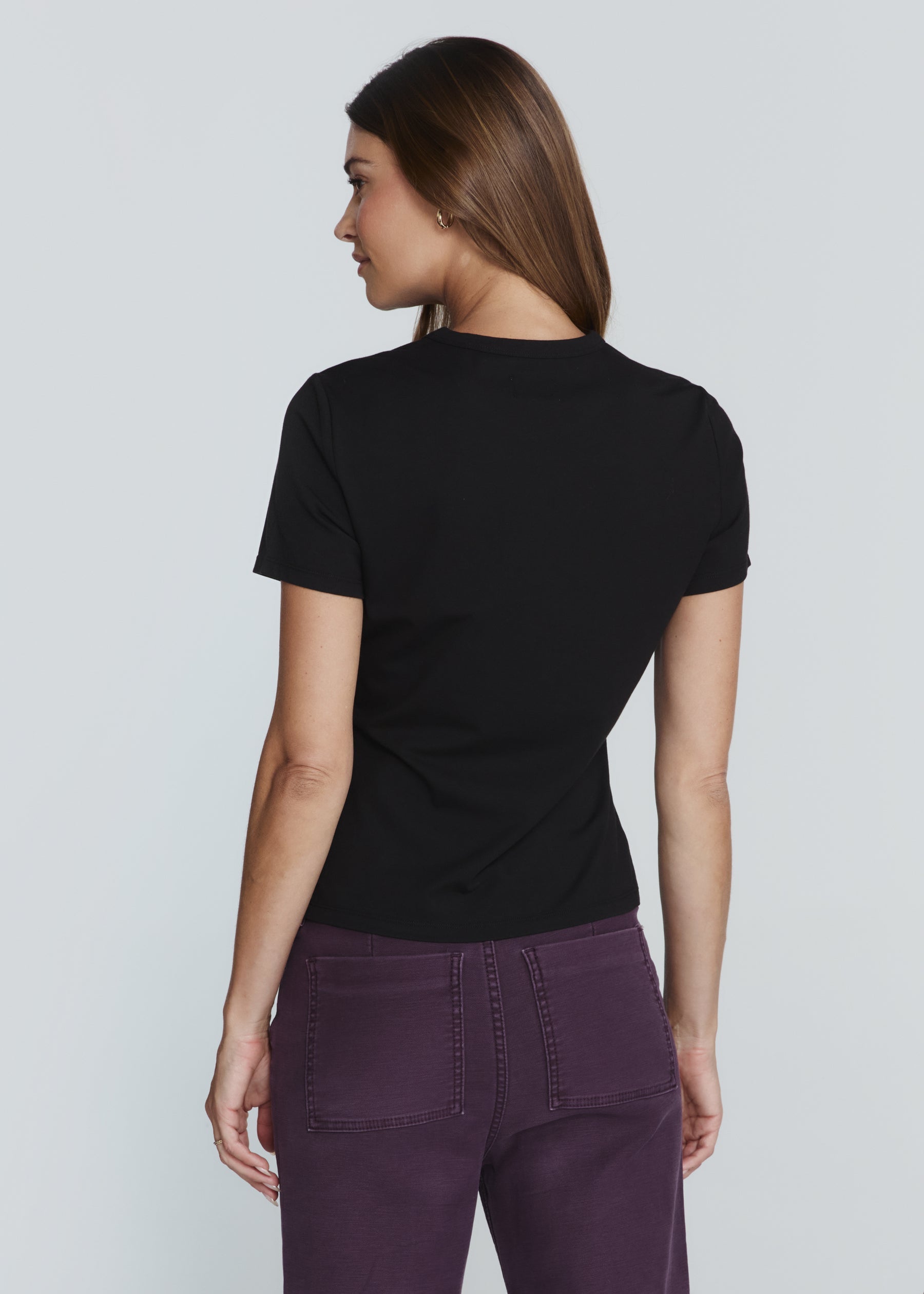 A woman shown from the back wearing a black short-sleeve crewneck t-shirt and purple pants, photographed against a solid light gray studio background.
