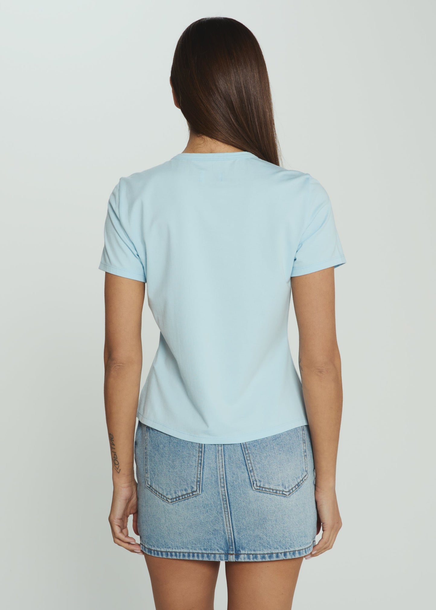 A woman viewed from the back wearing a light blue short-sleeve crewneck t-shirt and a light-wash denim mini skirt, set against a plain light gray studio background.