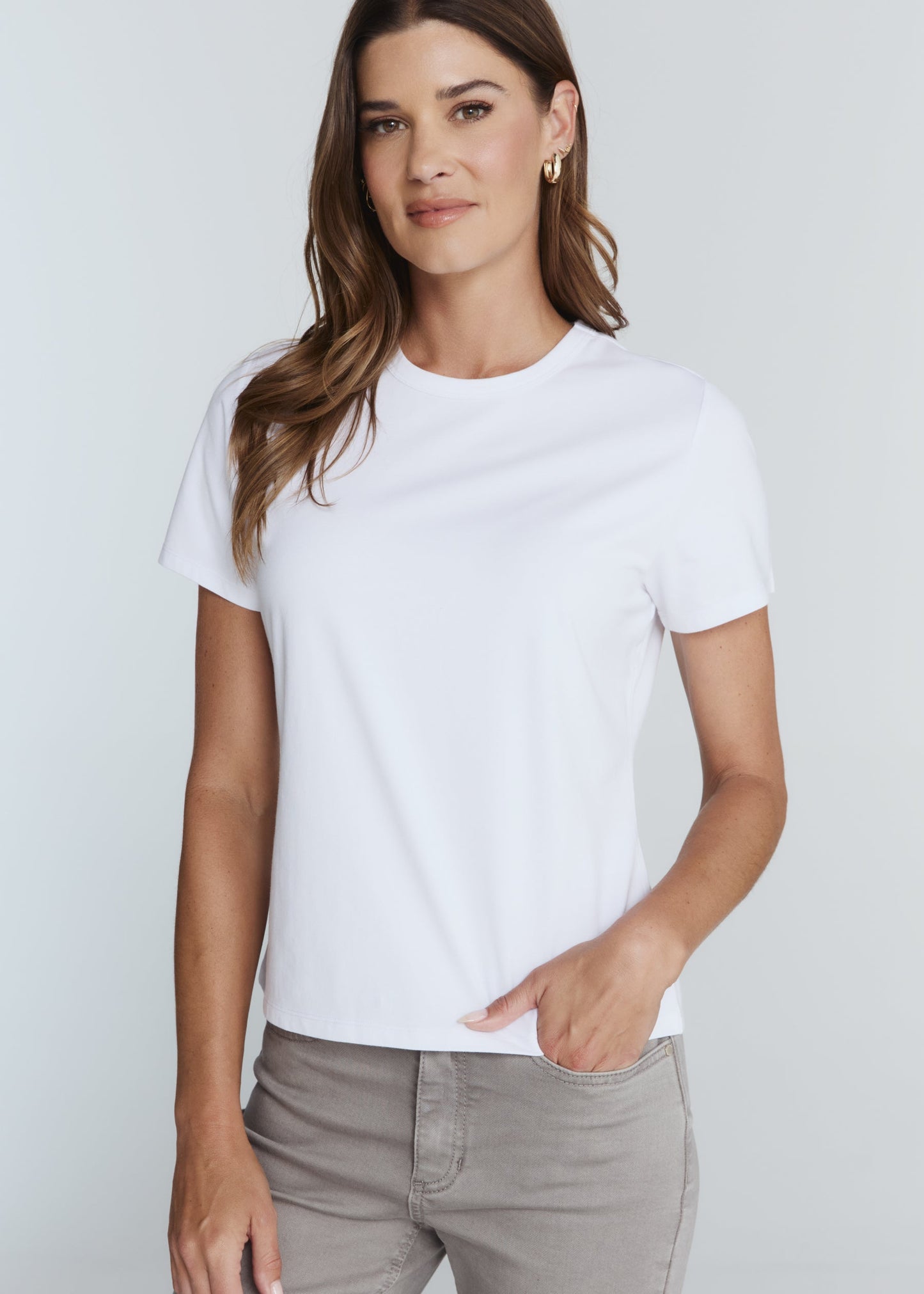 A woman wearing a plain white short-sleeve crewneck t-shirt and light gray pants, standing against a light gray studio background.