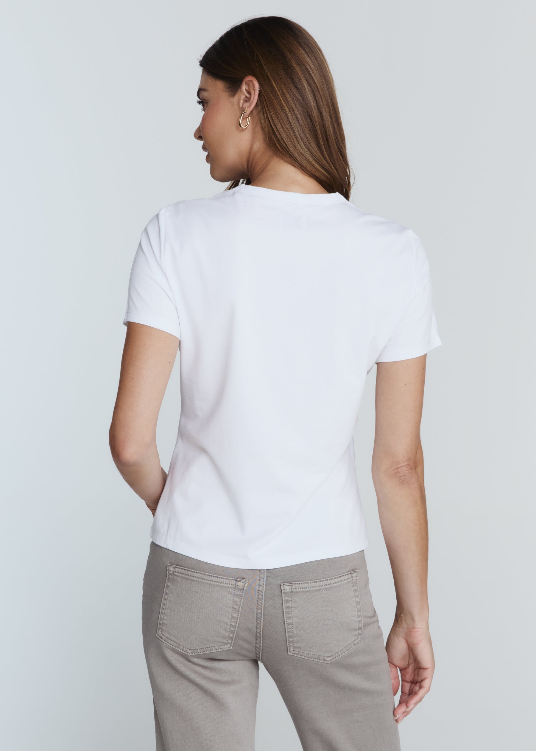 A woman shown from the back wearing a plain white short-sleeve crewneck T-shirt and light gray pants, photographed against a light gray studio background.
