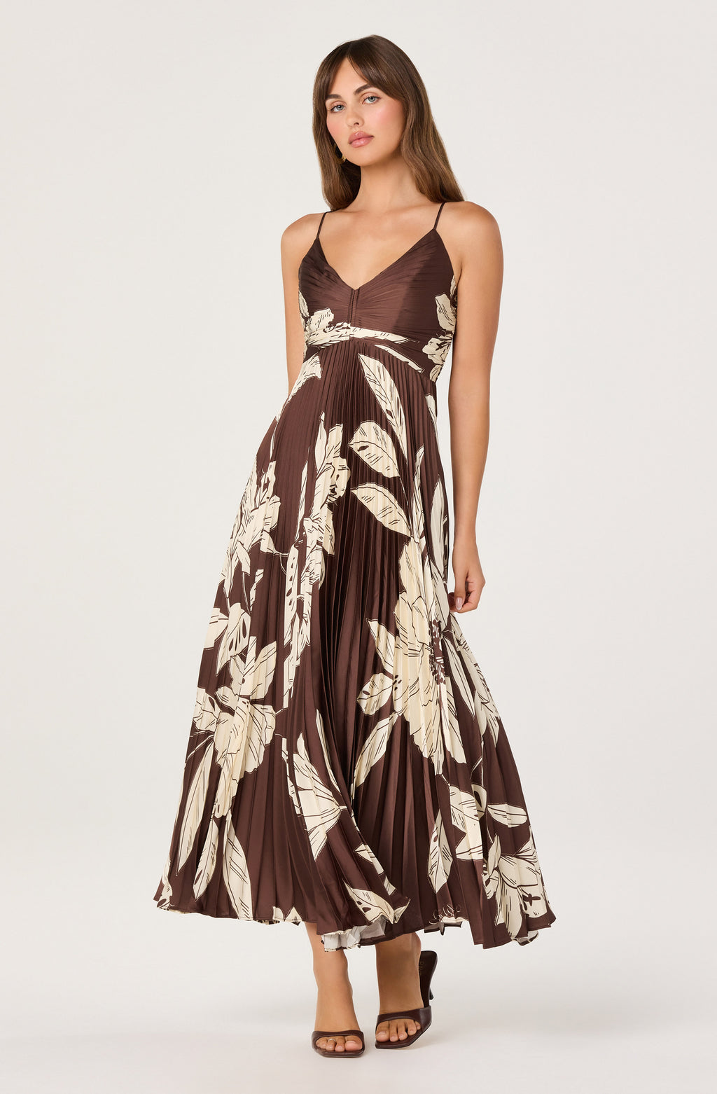 A woman modeling a brown maxi dress with a cream tropical leaf print, featuring spaghetti straps, a V-neckline, and a pleated skirt, set against a plain light gray background.