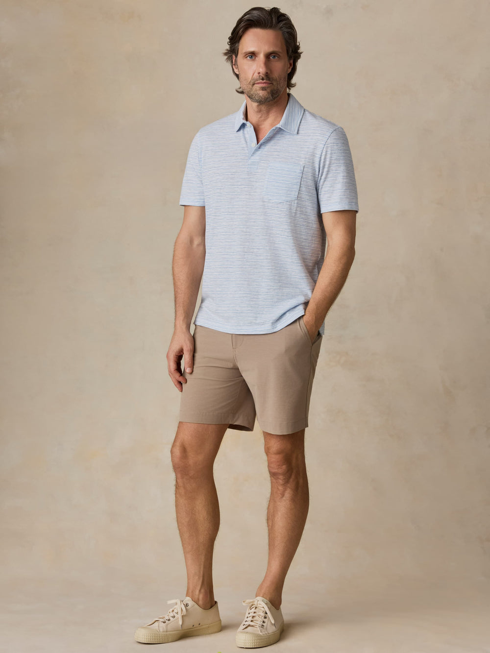 Man wearing a light blue striped polo shirt and beige shorts against a beige background