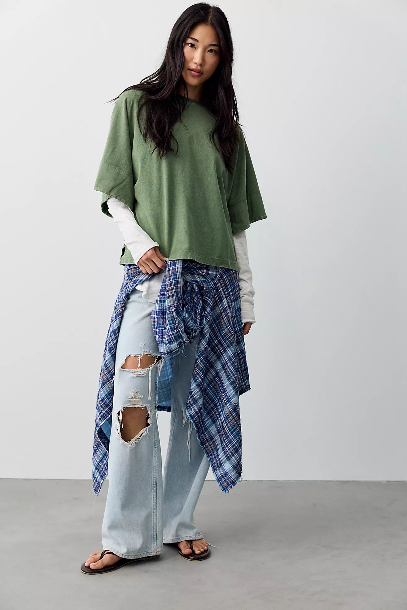 A woman modeling an oversized sage green t-shirt layered over a white long-sleeve shirt, paired with light-wash distressed jeans and a blue plaid flannel shirt tied around her waist, against a plain studio background.
