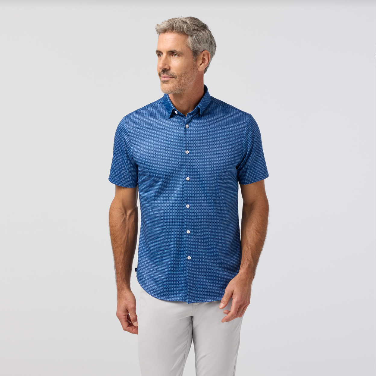 Man wearing a blue patterned shirt and white pants on a light gray background