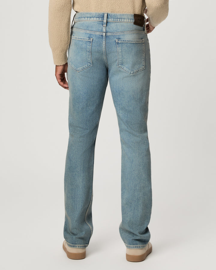 Rear view of a person wearing light-wash blue jeans with a beige sweater and tan shoes, standing against a plain light gray studio background.