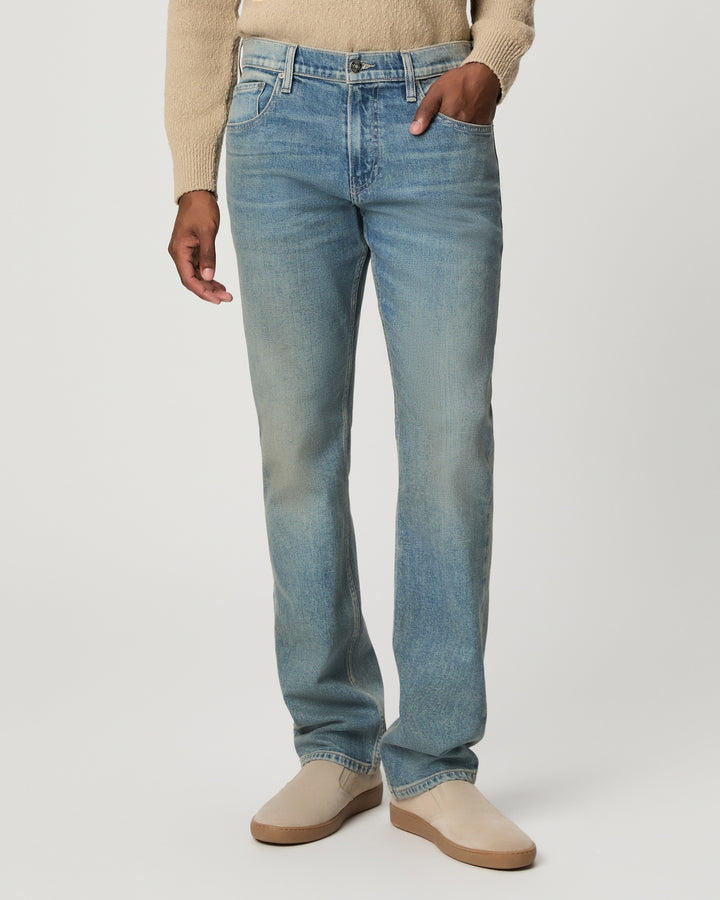 A person wearing light blue straight-leg denim jeans with a beige sweater and beige slip-on shoes, shown from the waist down against a light gray studio background.