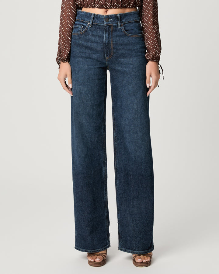 A person wearing dark blue wide-leg jeans with a brown patterned long-sleeve crop top and brown sandals, standing against a light gray studio background.