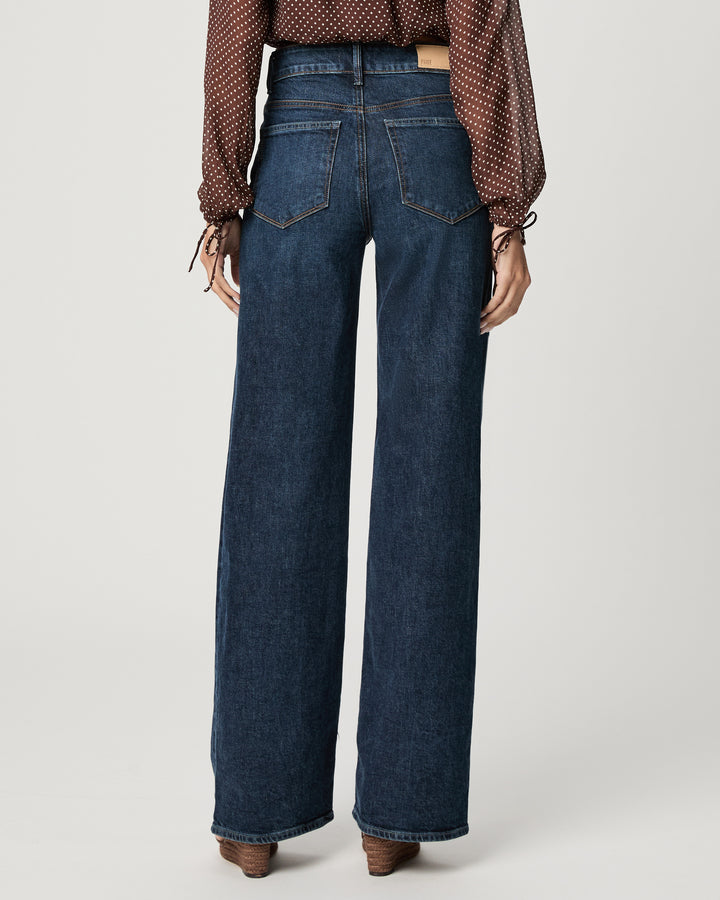 Rear view of a model wearing dark blue wide-leg denim jeans with a brown patterned blouse and brown shoes, set against a plain light gray studio background.