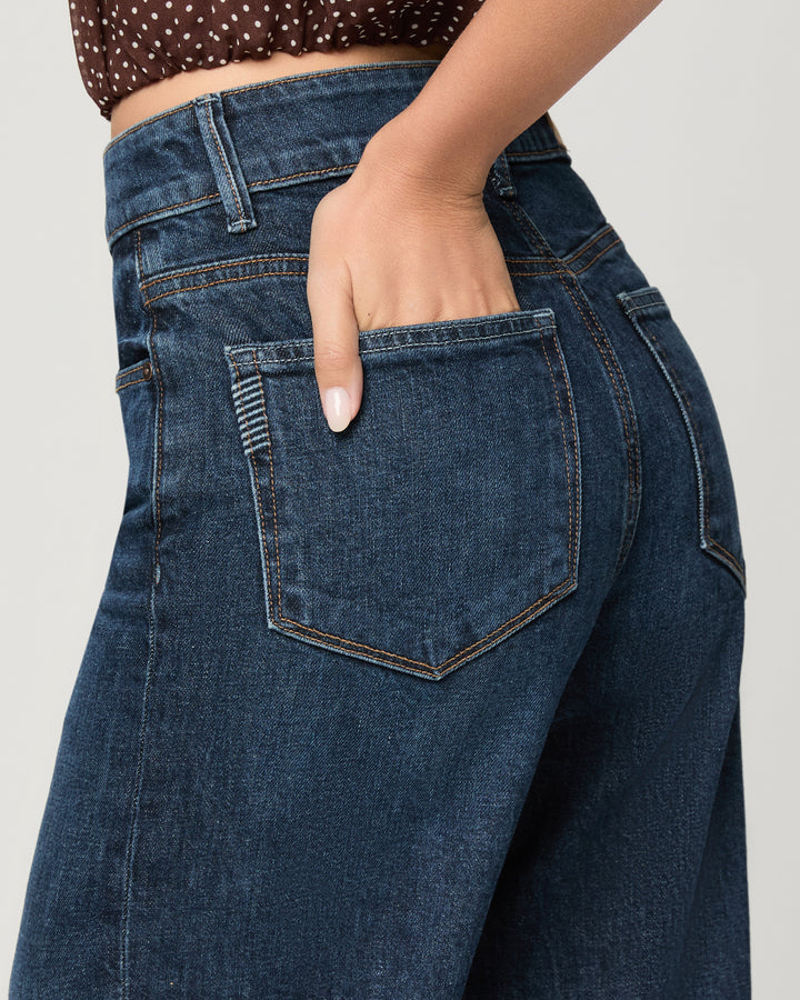 Close-up of a person wearing dark blue denim jeans, showing the back pocket, waistband, and hand in the pocket against a light gray studio background.
