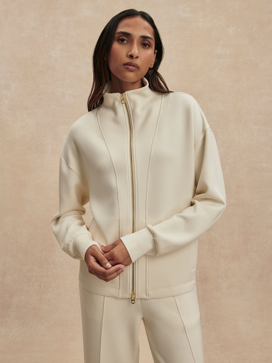 A woman wearing a cream-colored zip-up jacket and matching pants against a textured beige background.
