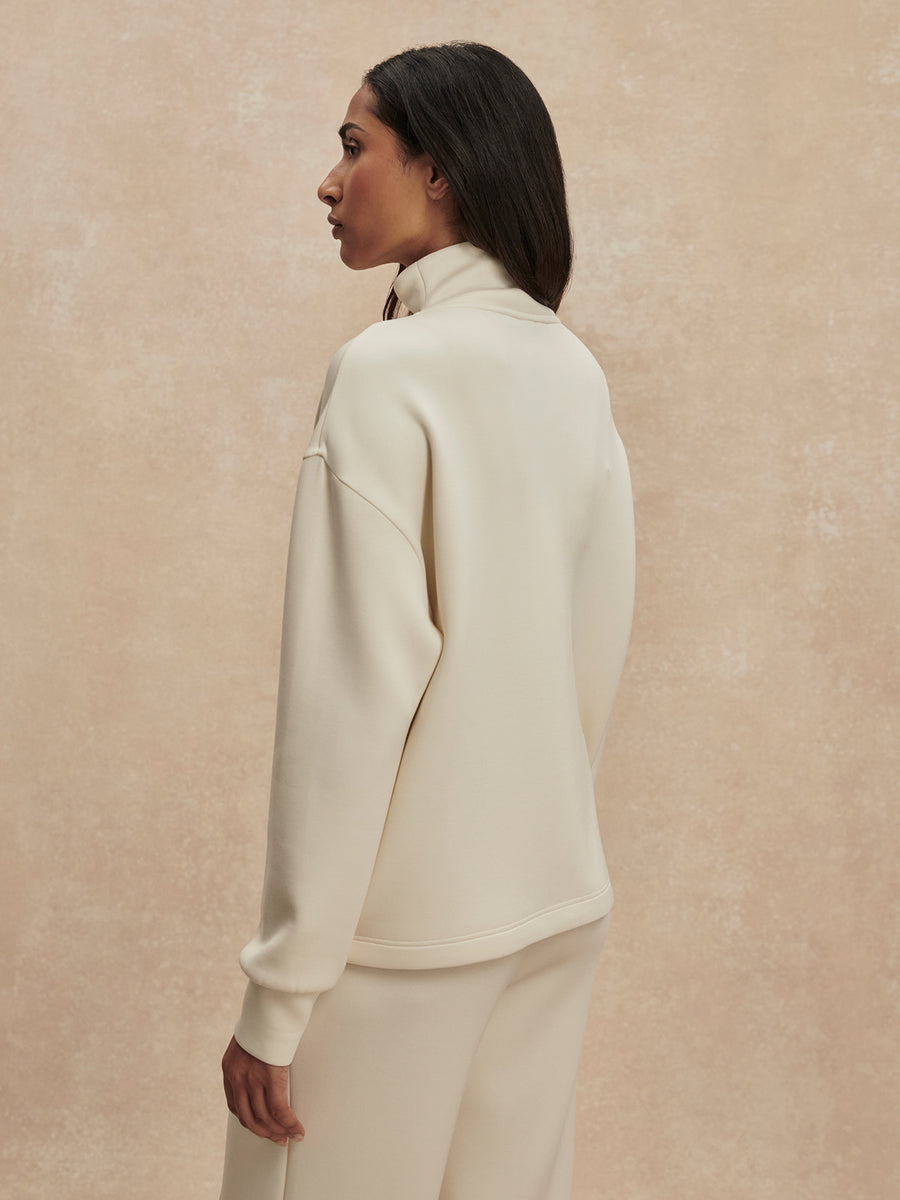 A woman shown from the back wearing a cream-colored mock-neck sweatshirt and matching sweatpants against a textured beige background.