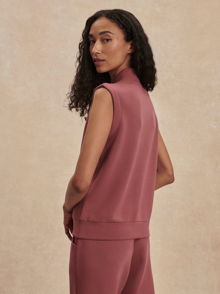 A woman wearing a sleeveless dusty rose vest and matching pants, photographed from the back against a textured beige studio backdrop.