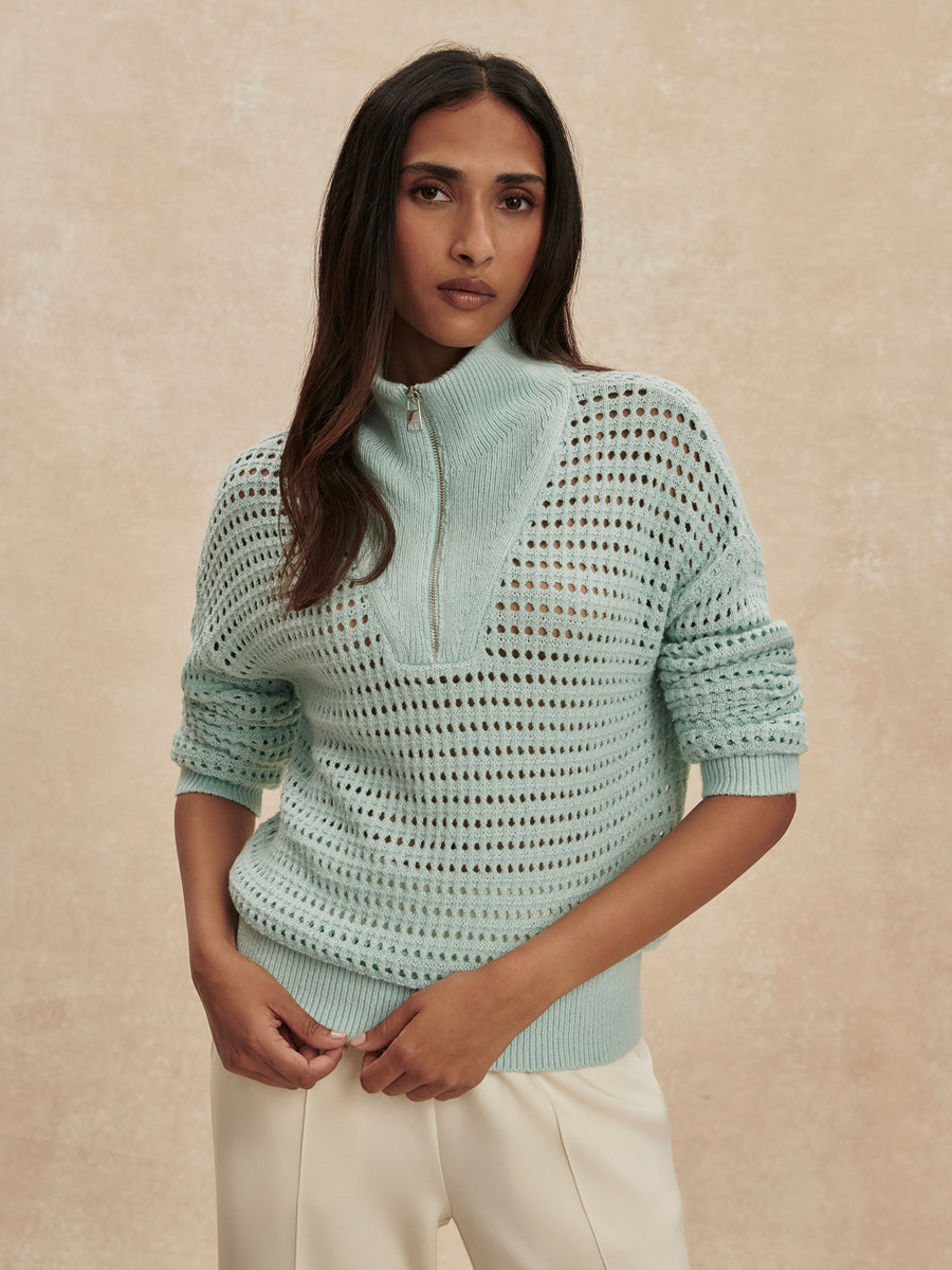 A woman wearing a pale mint green open-knit mock-neck sweater with a short zipper at the collar, paired with cream trousers, standing against a textured beige background.