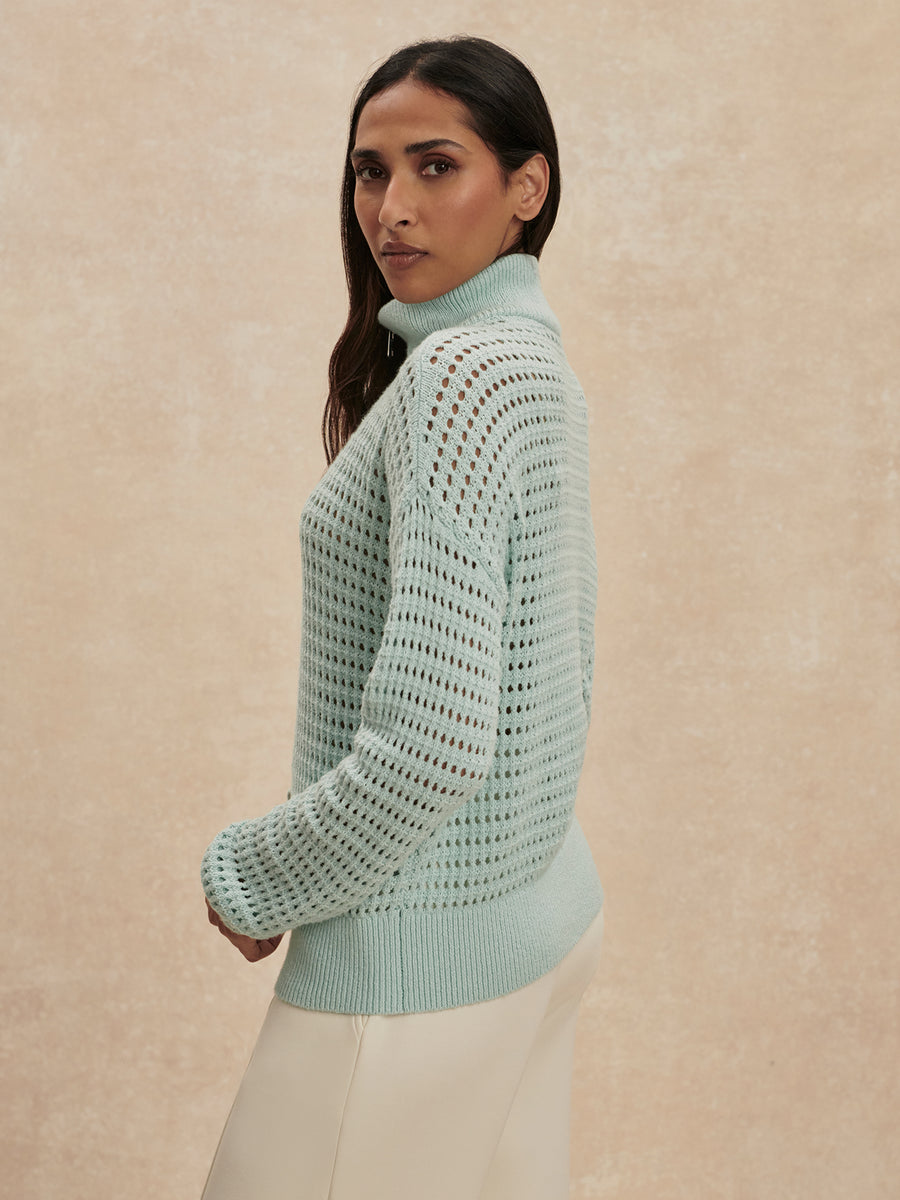A woman wearing a light mint green open-knit turtleneck sweater and cream pants, photographed against a textured beige studio backdrop.