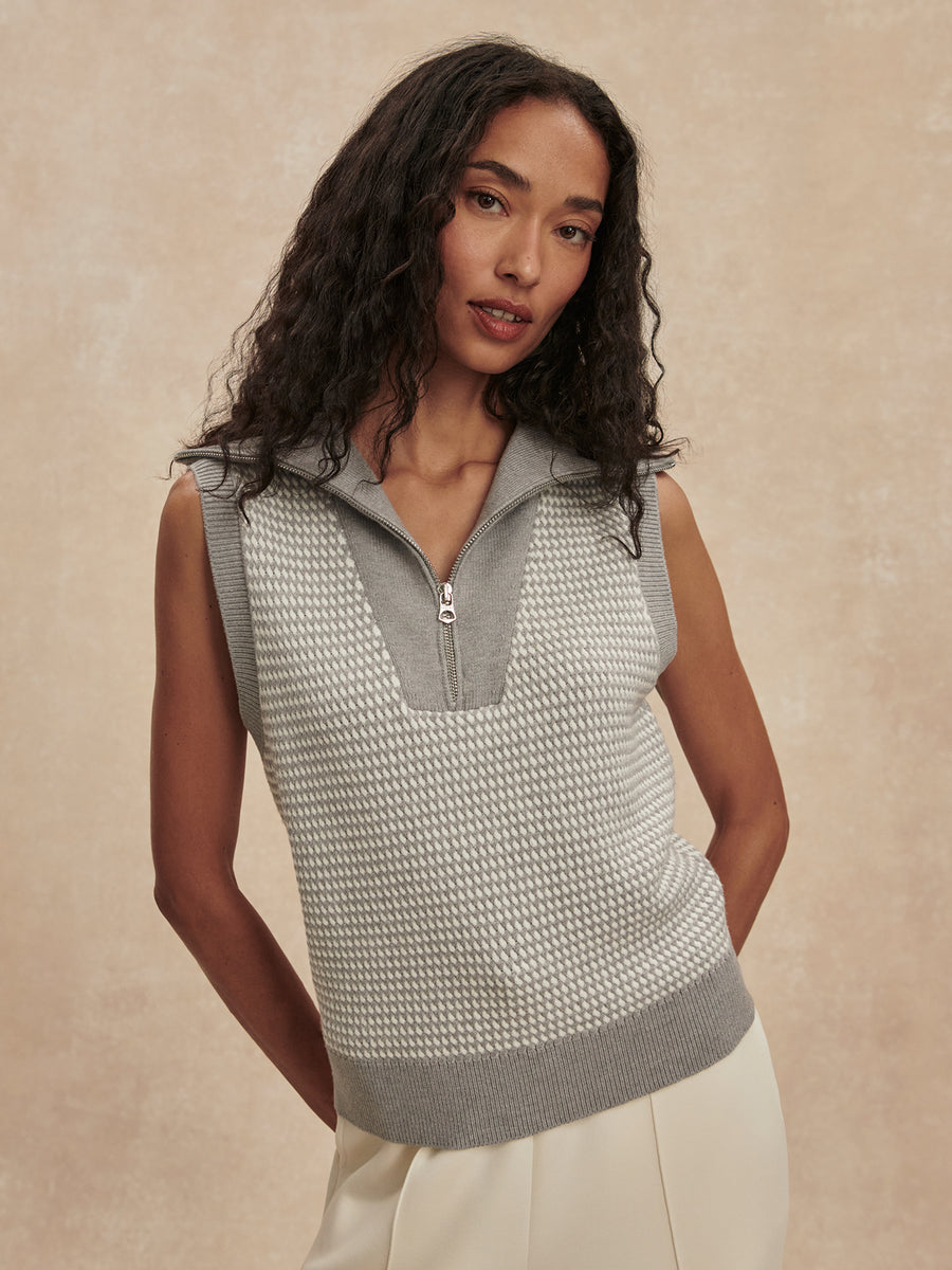 A woman wearing a sleeveless knit half-zip vest with a textured pattern in light gray and white, paired with cream-colored pants, set against a warm beige textured background.