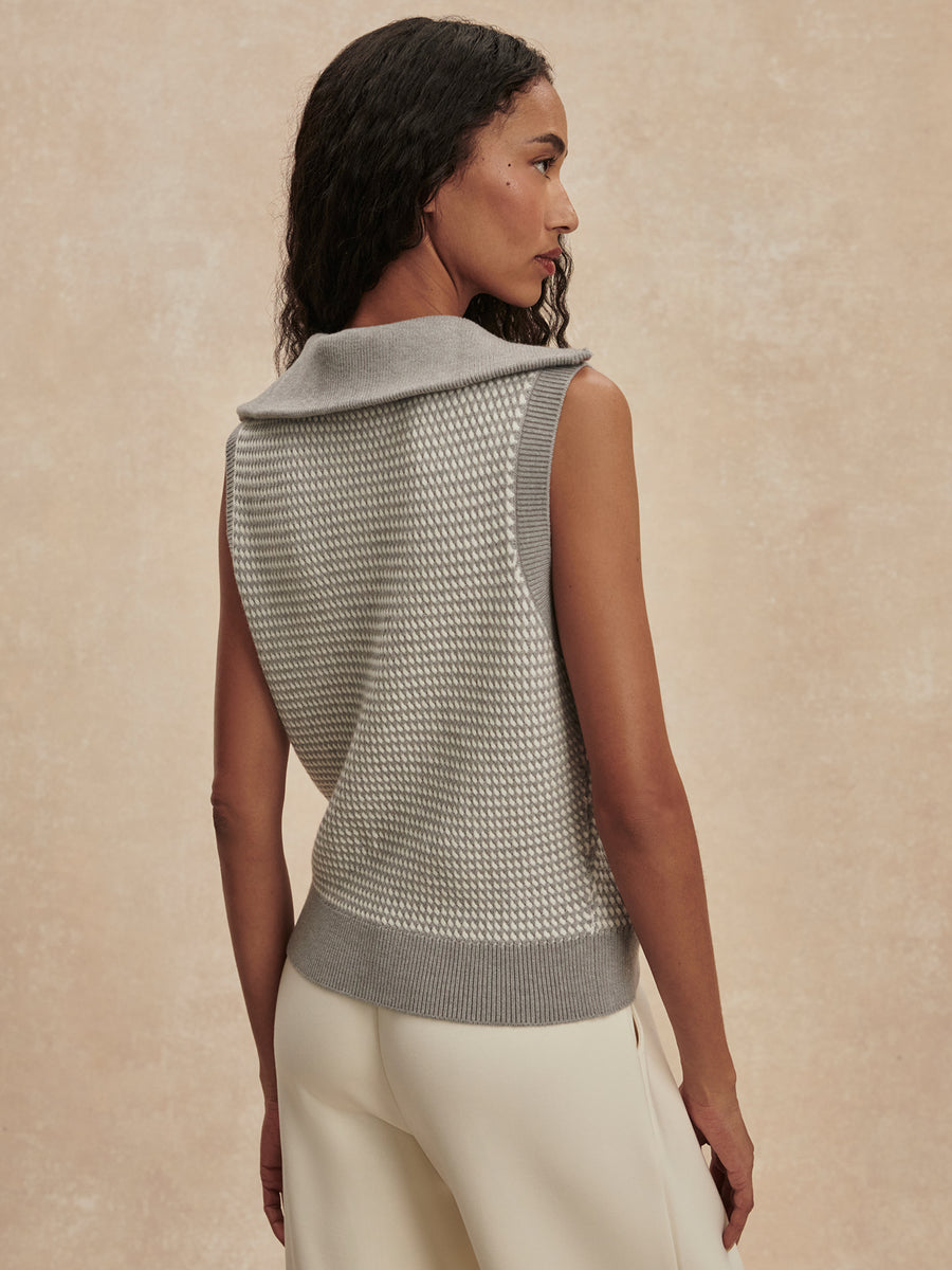 A woman shown from the back wearing a sleeveless knit vest with a wide folded collar and ribbed hem, paired with cream pants, against a warm beige textured studio background.