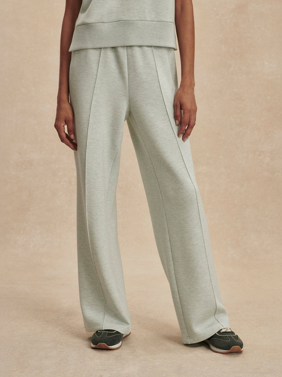 A model is shown from the waist down wearing light sage green wide-leg sweatpants, a matching top, and dark sneakers against a textured beige studio backdrop.