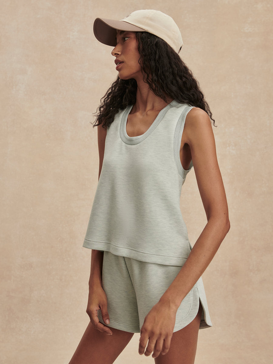 A woman wearing a light sage green sleeveless cropped tank top and matching shorts, paired with a beige baseball cap, posed against a textured beige studio background.